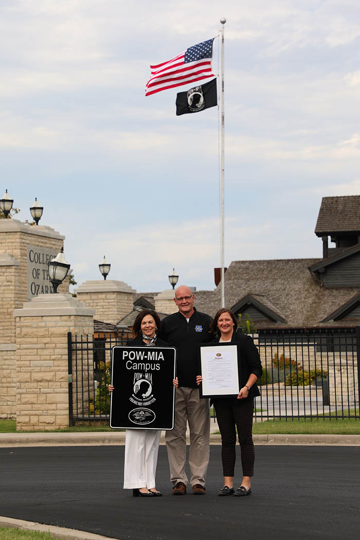 man and two women in front of flags holding POW MIA sign
